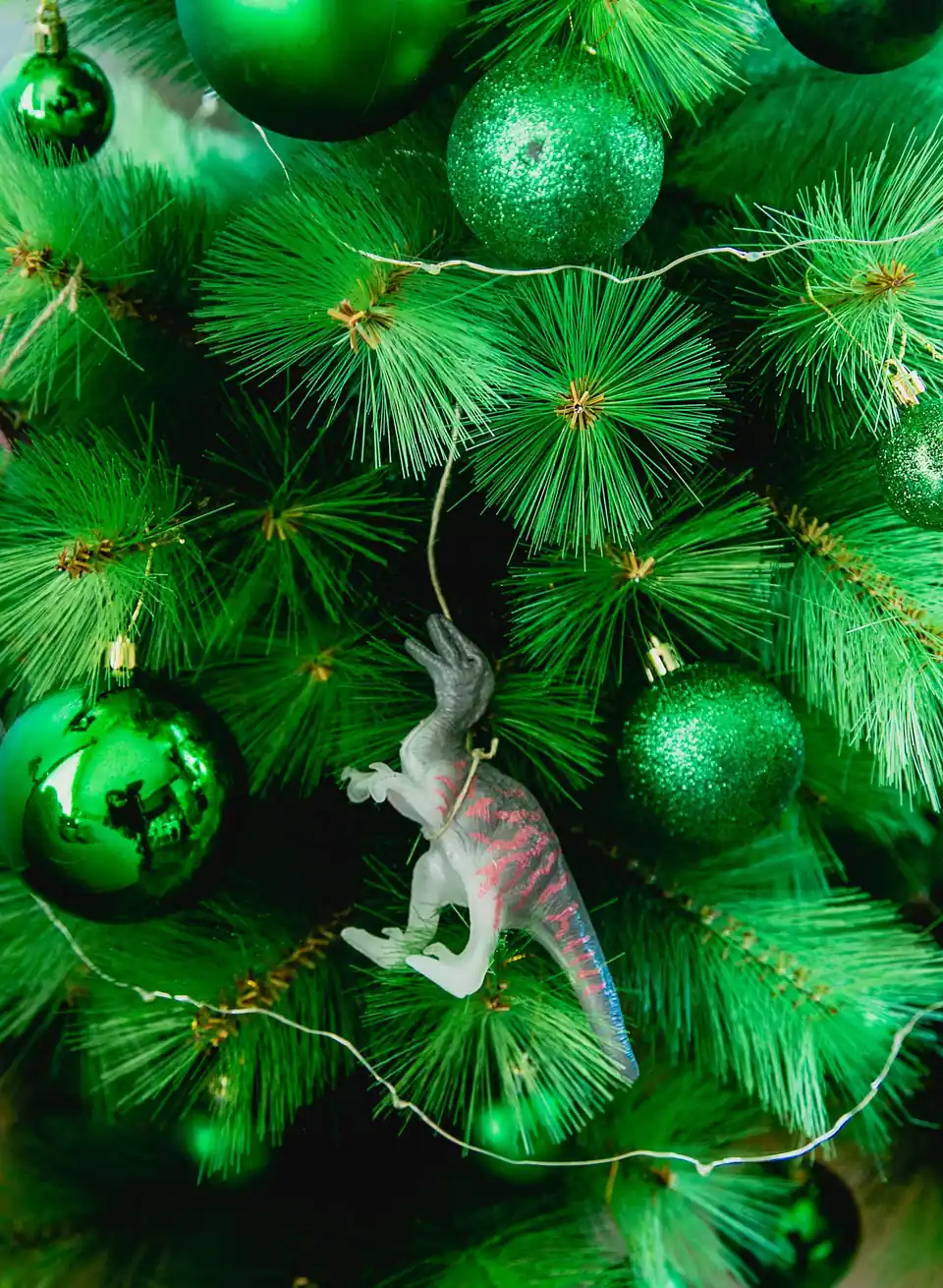 Close view of an artificial Christmas tree with long needle-style tips, green baubles, a thin wire light string and a white toy dinosaur ornament