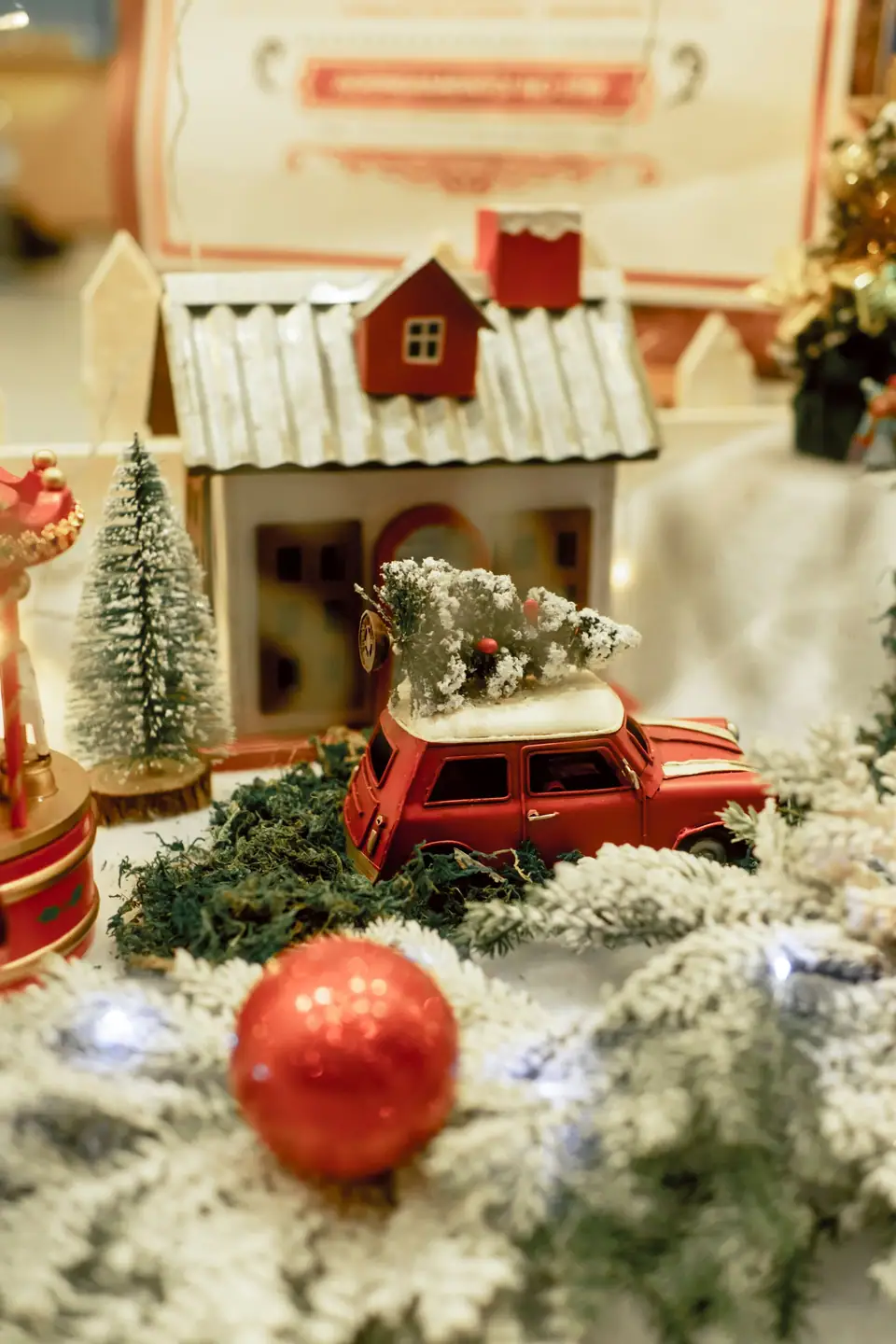 A small frosted bottle-brush tree beside a model house and a toy car carrying a tiny tree